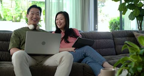 Asian couple sitting on sofa sharing laptop and tablet, working and relaxing at home