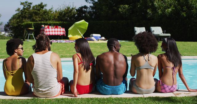 Young men and women of different ethnicities relaxing by pool in swimwear on a sunny day. Green lawn, trees, and a picnic setup with table and umbrella in background. Great for illustrating concepts related to summer fun, leisure, group activities, outdoor living, friendship and diversity in social settings.