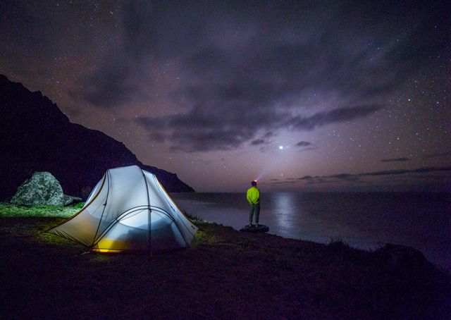 An individual camping near a mountain lake under a star-studded sky. The illuminated tent creates a serene contrast against the dark landscape. Ideal for use in outdoor adventure promotions, camping gear advertisements, or websites/blogs about hiking, stargazing, nature retreats, and solitude.