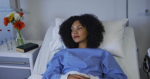 Calm female patient resting on hospital bed wearing blue gown looking thoughtful