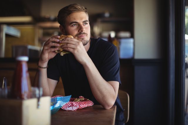 Young man sitting at a table in a restaurant, holding a burger and looking thoughtful. Ideal for use in advertisements for restaurants, fast food chains, or lifestyle blogs focusing on dining experiences and casual eating.