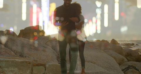 Couple embracing on rocky shore at dusk with city light bokeh double exposure
