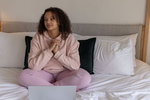 Woman sitting on bed using laptop and reflecting during cozy remote study session