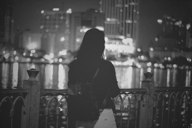 Silhouette of a woman with a backpack standing by a riverside at night, looking at a city skyline. The lights from the buildings create reflections on the water, adding a serene yet urban feel. Suitable for themes of introspection, urban lifestyle, night scenery, and peaceful moments in city life.
