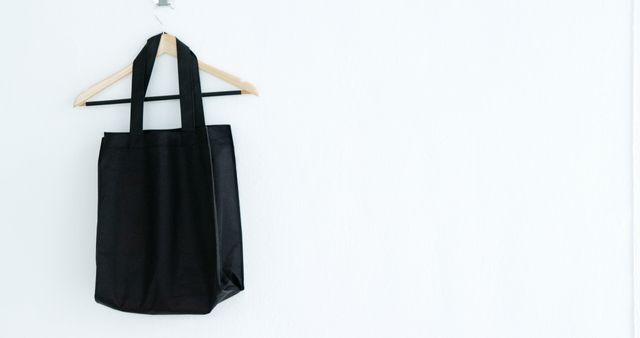 A black tote bag hangs on a wooden hanger against a white wall, with copy space. Its simplicity and the eco-friendly alternative it represents to plastic bags make it a popular accessory for shopping and personal use.