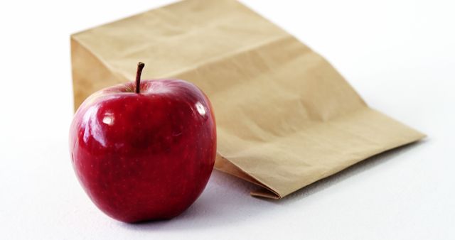 A shiny red apple sits in front of a brown paper bag on a white surface, with copy space. Often associated with health and education, the apple symbolizes healthy eating habits and is a common snack choice.