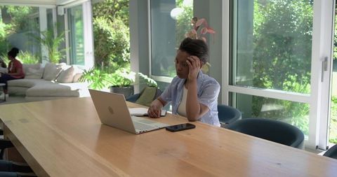 Young african woman working on laptop at bright home workspace with plants and sunlight