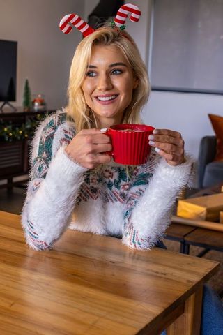 Smiling woman holding red mug in cozy holiday sweater with candy-cane headband