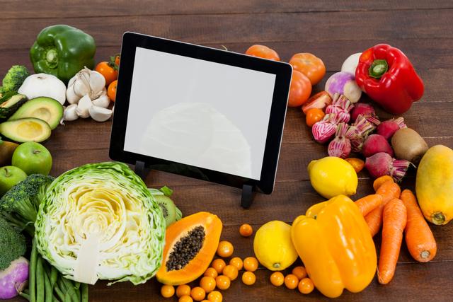 Digital tablet surrounded by an assortment of fresh vegetables and fruits on a wooden table. Ideal for use in articles or advertisements related to healthy eating, cooking, recipes, nutrition, and technology in the kitchen. Perfect for promoting organic produce, vegetarian or vegan lifestyles, and food preparation apps.