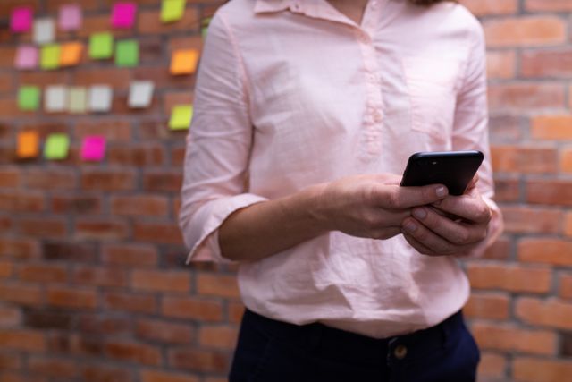Businesswoman using smartphone in a modern creative office with a brick wall background. Ideal for themes related to technology in the workplace, professional communication, modern work environments, and productivity.