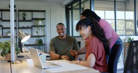 African American team collaborating and laughing at laptop in modern green office workspace