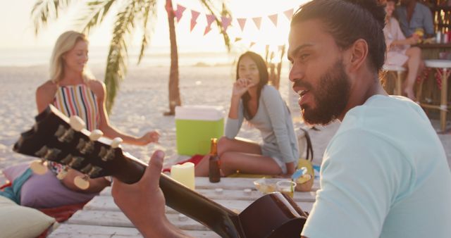 Young adults having a beach party during sunset, enjoying music with guitar, refreshments, and good company. Useful for travel brochures, summer event promotions, and lifestyle blogs related to outdoor activities and relaxation.