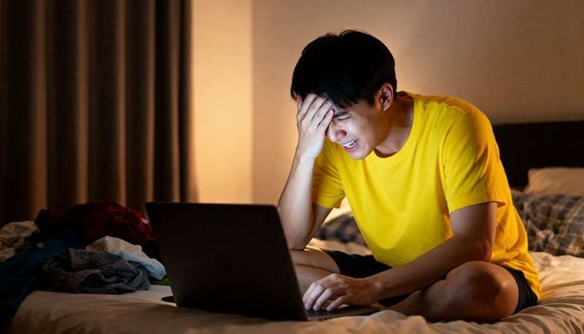 Frustrated young man pressing forehead while typing on laptop in warm bedroom night light