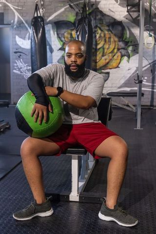 African american man holding green medicine ball sitting on bench in graffiti gym