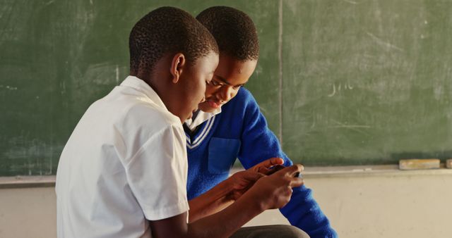Two African boys are keenly engaged with a smartphone in front of a classroom chalkboard, highlighting the intersection of technology with education. Useful for illustrating concepts related to modern education, elearning initiatives, or studies on digital literacy and childhood friendships.