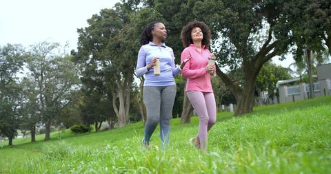 Diverse women walking together in park holding water bottles and smartphone conversation