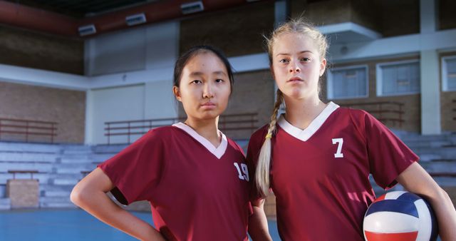 Two female volleyball players are standing in a gymnasium, dressed in red jerseys. The players appear determined and ready for their game, with one holding a volleyball. This image is ideal for depicting youth sports, teamwork, and gender diversity in athletics. It can be used for articles, advertisements, and promotional materials related to sports events, athletic programs, or team building activities.