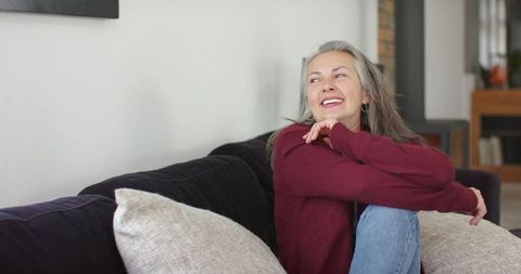 Smiling senior woman relaxing on sofa at home wearing burgundy sweater, cozy lifestyle
