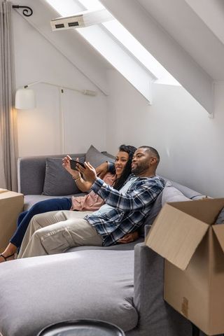 Couple relaxing on gray sofa in loft unpacking boxes and using smartphone under skylight