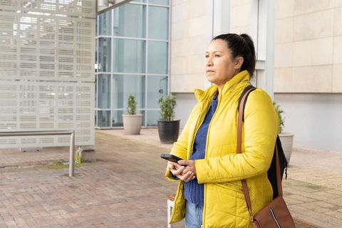 Mature woman standing in modern plaza wearing yellow puffer jacket holding smartphone