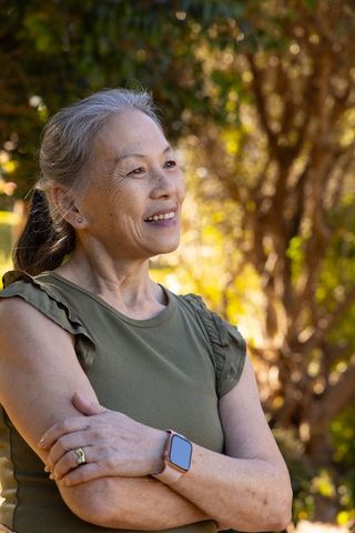 Senior asian woman smiling in sunlit garden wearing smartwatch and olive top