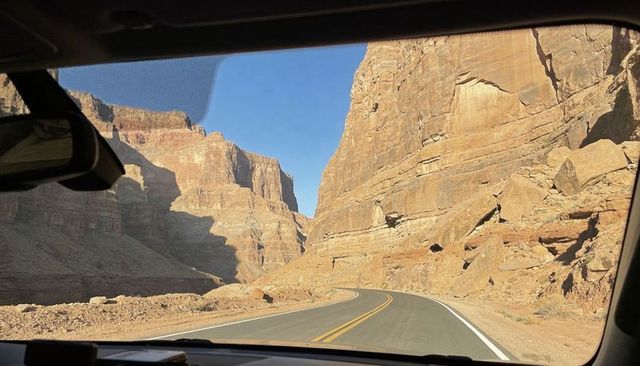 Driving through sunlit sandstone canyon on winding two-lane road framed by car windshield