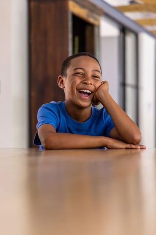 Laughing african boy leaning on wooden table in bright open-plan home, blue t-shirt
