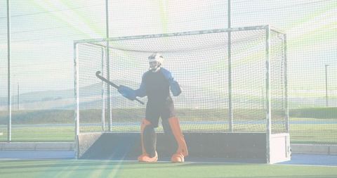 Standing field hockey goalkeeper wearing helmet and orange leg pads guarding outdoor net