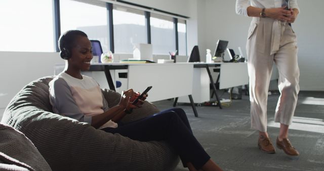 African american businesswoman wearing headphones, sitting in armchair while her coworker passing by. independent creative business at a modern office.