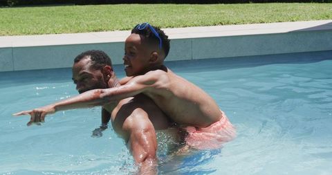 African father carrying son in backyard pool playing and pointing on sunny summer day