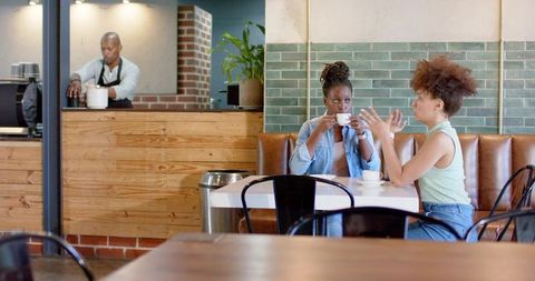 Two women chatting and sipping coffee in trendy cafe booth with barista and brick interior