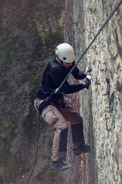 Individual scaling outdoor rock wall wearing helmet, harness, and climbing gear. Ideal for promoting adventure sports, outdoor recreation, safety equipment for climbing, or fitness activities involving physical endurance.