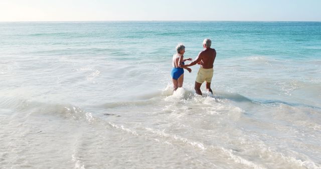 Senior couple enjoying themselves at the beach, splashing in the ocean water. Perfect for use in vacation advertisements, retirement planning materials, and lifestyle blogs.