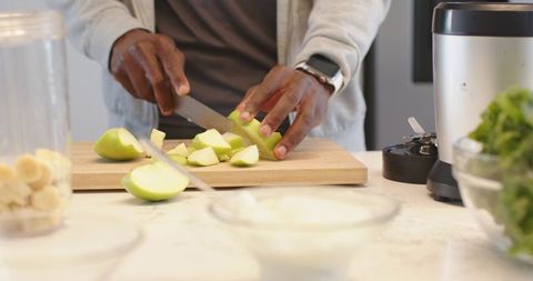 Hands slicing green apple on wooden board for smoothie prep with blender and smartwatch