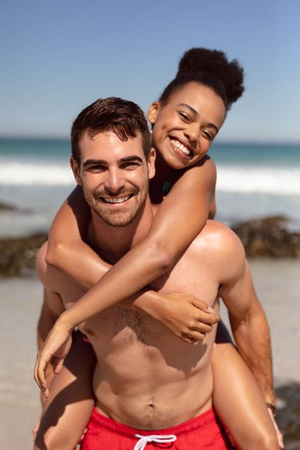 Front view of happy Caucasian man giving piggyback to woman on beach in the sunshine