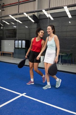 Diverse female friends walking on indoor paddle court holding paddles and laughing