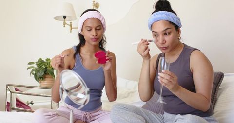 Two friends applying makeup on bed wearing plush headbands loungewear holding champagne glass