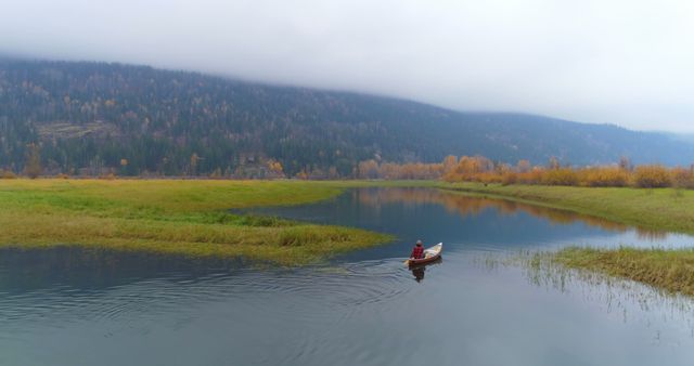 Serene landscape of a person canoeing on a calm lake surrounded by autumn foliage and mist-covered mountains. Water reflects surrounding nature creating a peaceful scene. Ideal for use in outdoor adventure promotions, travel blogs, nature conservation materials, and calming environment designs.