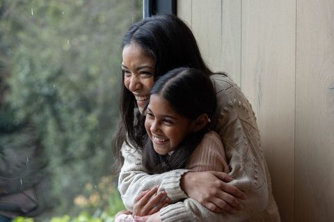 Indian mother hugging daughter on window seat during rainy day, cozy family moment