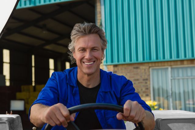 Worker in blue uniform driving tractor in industrial area, smiling confidently. Ideal for use in agricultural, industrial, and transportation-related content, showcasing job satisfaction and professional work environment.