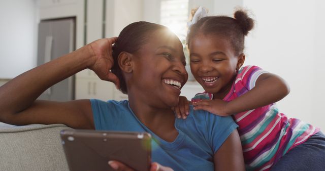 Happy african american mother and daughter sitting on sofa using digital tablet and laughing. staying at home in self isolation during quarantine lockdown.
