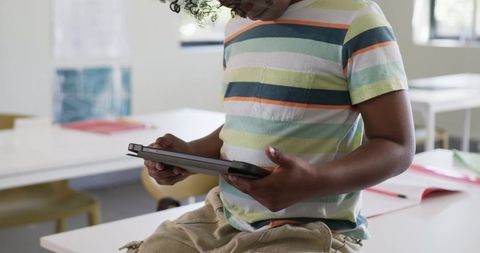 Young student using tablet in bright classroom wearing striped tee and casual khaki shorts