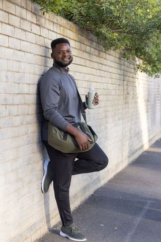 Smiling african man leaning on brick wall holding water bottle and duffel bag post workout