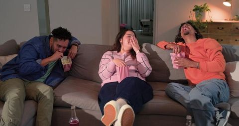 Friends laughing and eating popcorn on sofa during cozy movie night at home