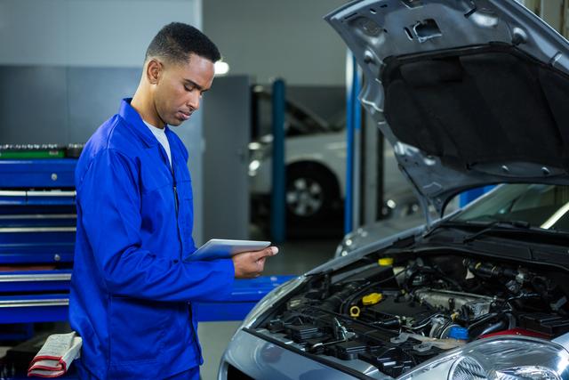 Mechanic in blue uniform using digital tablet while inspecting car engine in garage. Ideal for content related to automotive repair, modern technology in maintenance, professional services, and vehicle diagnostics.
