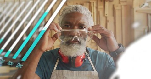 Senior craftsman raising safety goggles in woodworking shop wearing earmuffs and apron