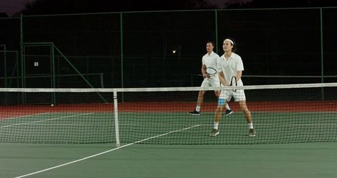 Two male tennis players playing doubles at net on outdoor hard court under night lighting