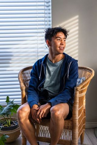 Asian teen sitting in rattan chair by window blinds, relaxed casual hoodie at home