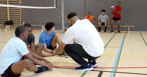 Diverse male volleyball teammates sitting and adjusting shoes on indoor gym court bench