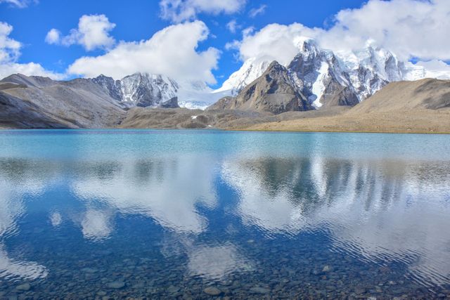 This beautiful scene shows a crystal-clear lake reflecting majestic snow-capped mountains and a bright blue sky. Ideal for nature, travel, or outdoor environmental themes. Perfect for use in articles about serene landscapes, clean water, and the beauty of high-altitude wilderness areas.